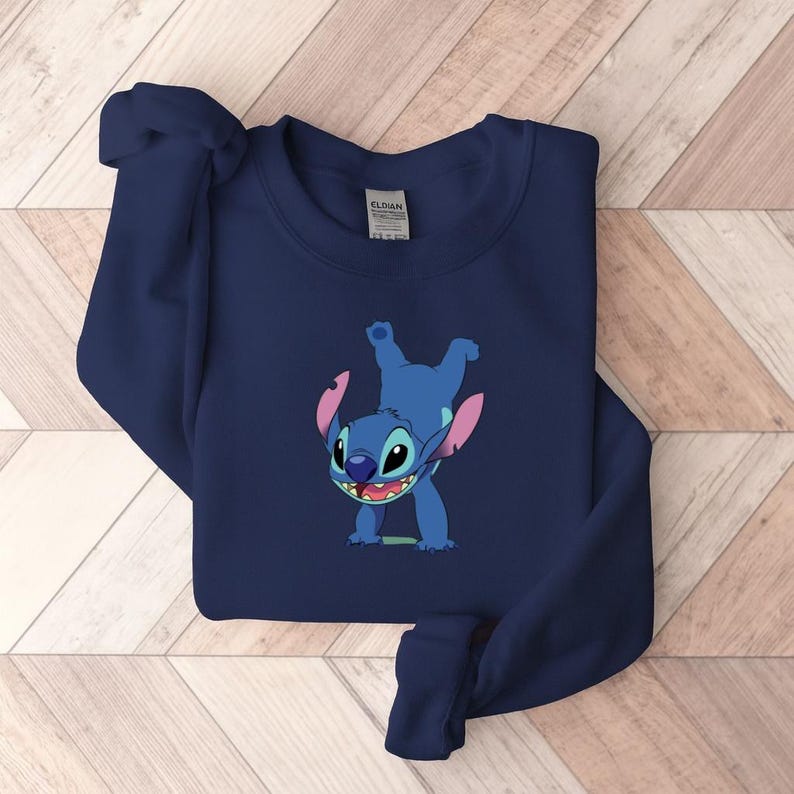 May include: Navy blue sweatshirt featuring a cartoon image of Stitch from Lilo & Stitch. The blue character is in a playful pose with his arms and legs in the air. The sweatshirt is made of a soft material.