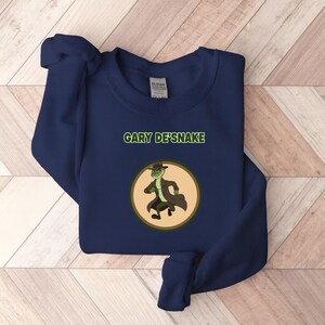 May include: A navy blue sweatshirt with the text "GARY DE'SNAKE" above a circular graphic. The graphic shows a cartoon snake wearing a fedora and trench coat. The sweatshirt is made of a soft material.