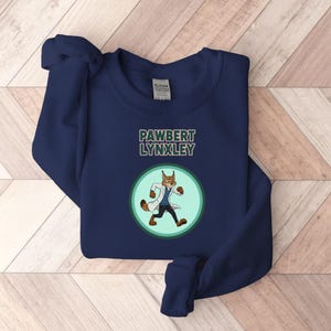 May include: Navy blue sweatshirt with the text "PAWBERT LYNXLEY" above a cartoon lynx wearing a lab coat. The lynx is inside a green and light green circle. The sweatshirt is made of a soft material.