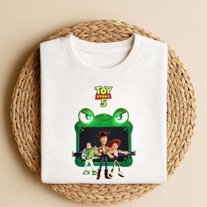 May include: White t-shirt featuring a graphic of characters from Toy Story 5. The design includes Woody, Buzz Lightyear, and Jessie, set against a green frog-shaped background. The shirt has a crew neck.