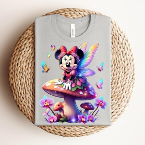 May include: A light gray t-shirt featuring a colorful illustration of Minnie Mouse as a fairy, sitting on a mushroom. The design includes butterflies and flowers. The shirt is displayed on a woven, circular placemat.