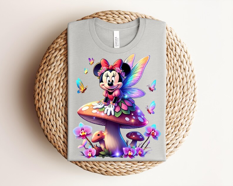 May include: A light gray t-shirt featuring a colorful illustration of Minnie Mouse as a fairy, sitting on a mushroom. The design includes butterflies and flowers. The shirt is displayed on a woven, circular placemat.