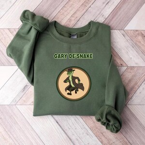 May include: An olive green sweatshirt with the text "GARY DE'SNAKE" above a circular graphic. The graphic shows a cartoon snake in a trench coat and fedora. The sweatshirt is made of a soft material.