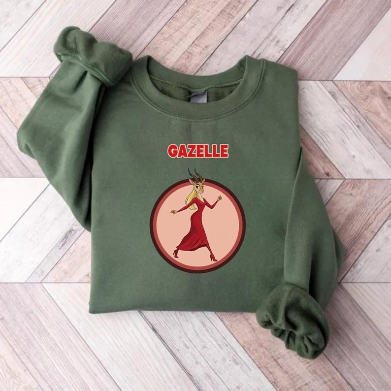 May include: An olive green sweatshirt with the word "GAZELLE" in red above a circular graphic. The graphic shows a cartoon gazelle in a red dress. The sweatshirt is made of a soft material.