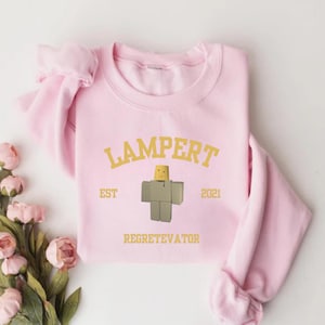 May include: A light pink sweatshirt with the word "LAMPERT" in gold lettering above a block figure. The text "EST 2021" is on either side of the figure, and "REGRETEVATOR" is below.
