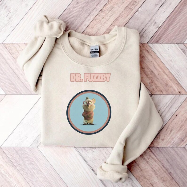 May include: A cream-coloured sweatshirt displaying the text "DR. FUZZBY" above a circular graphic. The graphic features a cartoon bear wearing glasses and a sweater, set against a light blue background with a brown and black border. The sweatshirt is folded on a wooden surface.