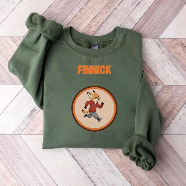Op de afbeelding: Een olijfgroene sweatshirt met de naam "FINNICK" in oranje blokletters boven een cirkelvormige afbeelding. De afbeelding toont een cartoonvos in een rode jas en grijze broek die rent. De sweatshirt is op een houten oppervlak gevouwen.