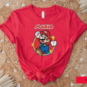 Może przedstawiać: Czerwona koszulka z grafiką postaci Mario z gry wideo Super Mario Bros. Projekt zawiera imię Mario w białej i czerwonej czcionce, z Mario w jego charakterystycznym stroju. Koszulka jest wykonana z miękkiego materiału.