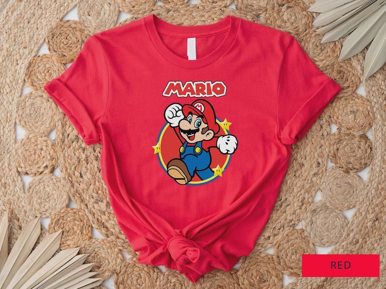 Może przedstawiać: Czerwona koszulka z grafiką postaci Mario z gry wideo Super Mario Bros. Projekt zawiera imię Mario w białej i czerwonej czcionce, z Mario w jego charakterystycznym stroju. Koszulka jest wykonana z miękkiego materiału.