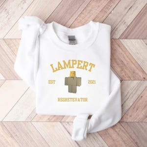 May include: White sweatshirt with the word "LAMPERT" in gold lettering. Below the word "LAMPERT" is a cartoon character and the words "EST 2021" and "REGRETEVATOR" in gold lettering. The sweatshirt is on a wooden background.