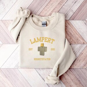 May include: A cream-colored sweatshirt with the word "LAMPERT" arched across the top in gold lettering. Below the word is a graphic of a block figure with a yellow head. The text "EST 2021" and "REGETEVATOR" are also printed on the sweatshirt.