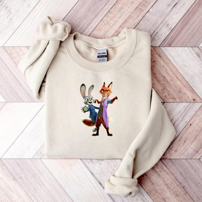 May include: A cream-colored sweatshirt featuring a graphic of the animated characters Judy Hopps and Nick Wilde from Zootopia. Judy, a gray rabbit, wears a blue jacket, while Nick, a fox, is in a purple coat. The sweatshirt is folded on a wooden surface.