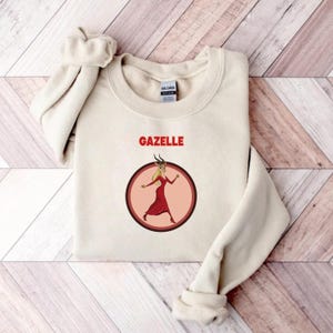 May include: A cream-coloured sweatshirt with the word "GAZELLE" in red block letters above a circular graphic. The graphic features a cartoon woman in a red dress with horns, inside a brown and pink circle.