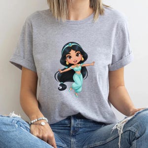 Puede incluir: Camiseta gris con una ilustraci&oacute;n de dibujos animados de una princesa con un atuendo turquesa y pelo largo y negro. La princesa sonr&iacute;e con los brazos extendidos. La camiseta tiene las mangas enrolladas.