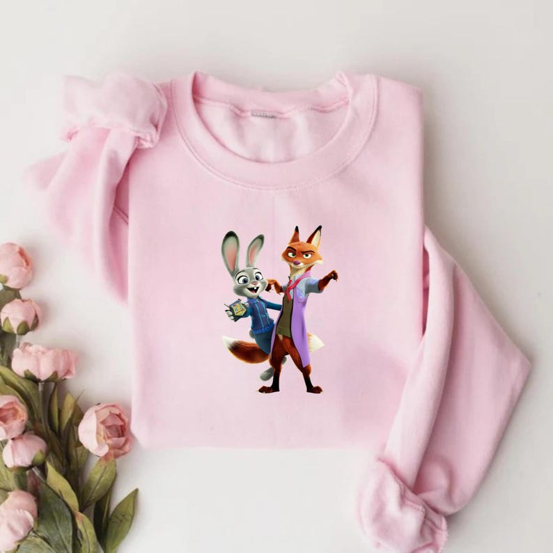 May include: A light pink sweatshirt featuring a cartoon image of Judy Hopps and Nick Wilde from Zootopia. Judy is a gray rabbit wearing a blue uniform, and Nick is a fox in a purple coat. The sweatshirt has long sleeves.
