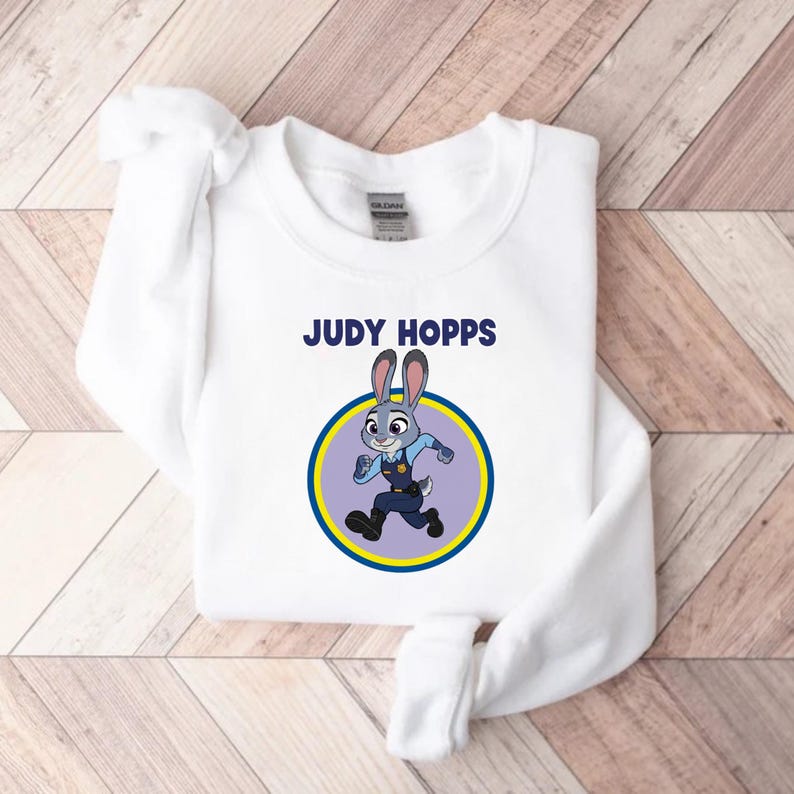 以下が含まれることがあります： 「JUDY HOPPS」の文字と、警察官の制服を着たウサギの漫画のイラストが描かれた白いスウェットシャツ。 ウサギは黄色と紫の円の中に入っています。
