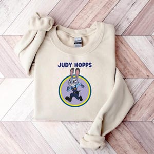 以下が含まれることがあります： 「JUDY HOPPS」の文字と、警察官の制服を着たウサギのキャラクター、ジュディ・ホップスの漫画のイメージが、青と黄色の円の中にあるクリーム色のスウェットシャツ。スウェットシャツは木製の表面に折りたたまれています。