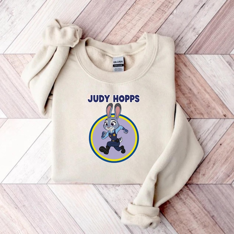 以下が含まれることがあります： 「JUDY HOPPS」の文字と、警察官の制服を着たウサギのキャラクター、ジュディ・ホップスの漫画のイメージが、青と黄色の円の中にあるクリーム色のスウェットシャツ。スウェットシャツは木製の表面に折りたたまれています。