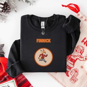 Op de afbeelding: Zwart sweatshirt met de naam "FINNICK" in oranje letters. Daaronder een ronde afbeelding van een cartoonvos met een rode jas en een grijze broek. De vos rent. Een zilveren ketting is zichtbaar.