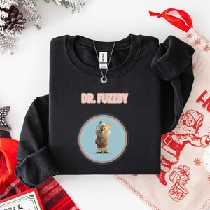 May include: Black sweatshirt with pink text reading "DR. FUZZBY". The sweatshirt displays a circular graphic of a cartoon character wearing glasses and a sweater. A silver necklace with a sun pendant is visible.