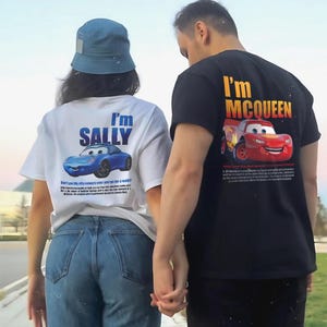 May include: White t-shirt with a blue cartoon car and the text "I'm SALLY". Black t-shirt with a red cartoon car and the text "I'm McQUEEN". The shirts are worn by two people holding hands.