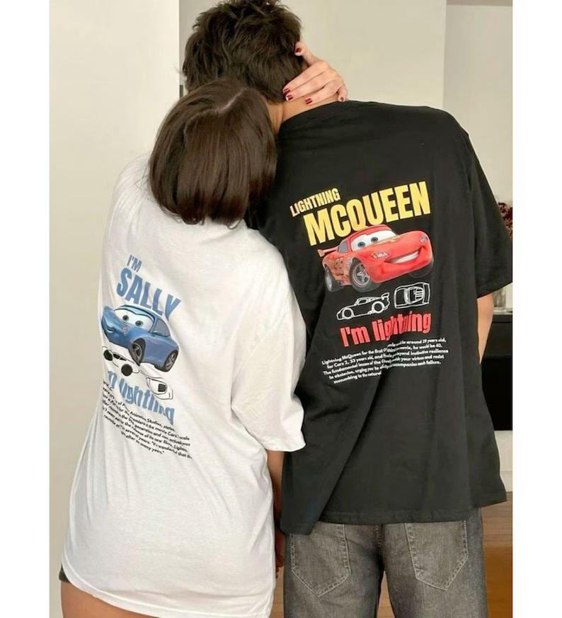May include: Two people wearing matching graphic t-shirts. One shirt is white with a blue cartoon car and the text "I'm Sally". The other shirt is black with a red cartoon car and the text "Lightning McQueen".