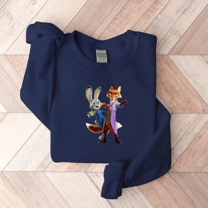 May include: Navy blue sweatshirt featuring a colorful graphic of the characters Judy Hopps and Nick Wilde from the animated movie Zootopia. The sweatshirt is made of a soft material and has a crew neck.