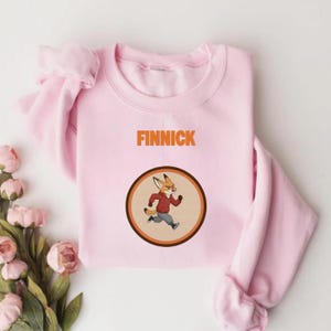 Op de afbeelding: Een lichtroze sweatshirt met het woord "FINNICK" in oranje letters. Daaronder een cirkelvormige afbeelding van een rennend vos-personage met een rode jas en grijze broek. De sweatshirt is gemaakt van zacht materiaal.