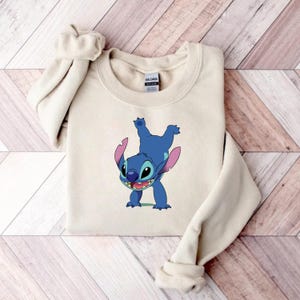 May include: A beige sweatshirt featuring a blue cartoon character, Stitch, in a playful pose. The character has large ears, a wide smile, and is depicted upside down. The sweatshirt is made of a soft material.