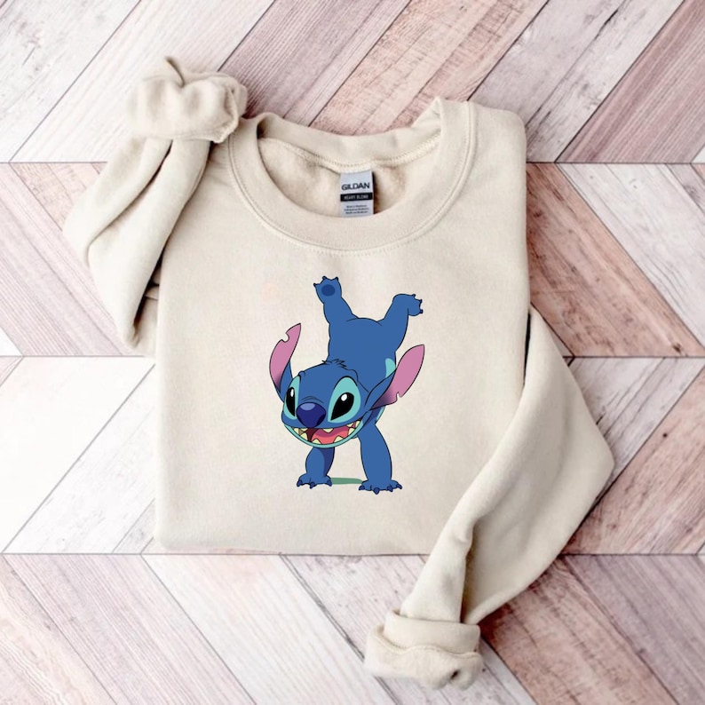 May include: A beige sweatshirt featuring a blue cartoon character, Stitch, in a playful pose. The character has large ears, a wide smile, and is depicted upside down. The sweatshirt is made of a soft material.