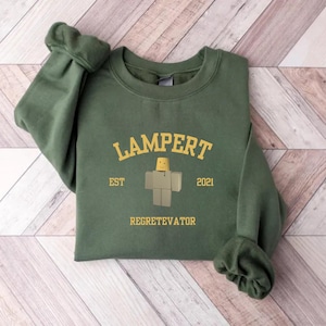 May include: Green sweatshirt with the word "LAMPERT" in gold lettering above a block figure and the words "EST 2021" on either side. Below the figure is the word "REGETEVATOR". The sweatshirt is folded on a wooden surface.