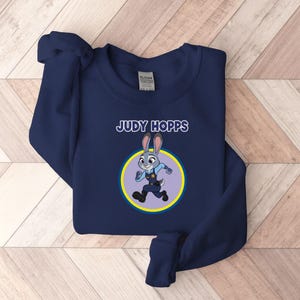 以下が含まれることがあります： 「JUDY HOPPS」の文字と、ウサギのキャラクター、ジュディ・ホップスの漫画のイメージが描かれたネイビーブルーのスウェットシャツ。 画像は黄色と青の円の中にあります。 スウェットシャツは柔らかい素材で作られています。