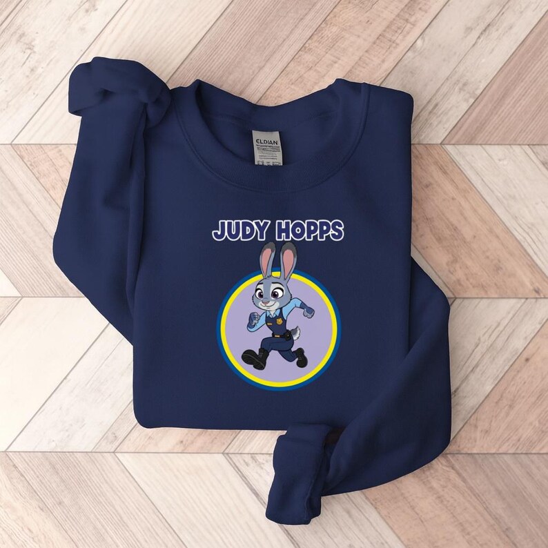 以下が含まれることがあります： 「JUDY HOPPS」の文字と、ウサギのキャラクター、ジュディ・ホップスの漫画のイメージが描かれたネイビーブルーのスウェットシャツ。 画像は黄色と青の円の中にあります。 スウェットシャツは柔らかい素材で作られています。