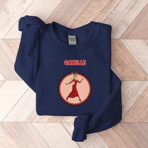 May include: A navy blue sweatshirt featuring the word "GAZELLE" in red above a cartoon gazelle in a red dress, within a pink circle. The gazelle has blonde hair and is wearing red heels.