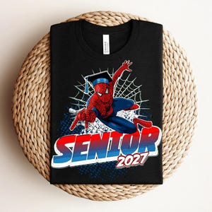 May include: A black t-shirt featuring a graphic of a red and blue superhero wearing a graduation cap and shooting a web. Below the character, the word SENIOR is printed in large red and blue letters, followed by the year 2027 in white text.