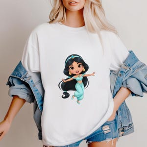 Puede incluir: Camiseta blanca con una ilustraci&oacute;n de dibujos animados de una princesa con un atuendo turquesa, pelo largo negro, una diadema azul y pendientes dorados. La princesa est&aacute; en una pose de baile. La camisa la lleva una persona con una chaqueta vaquera.