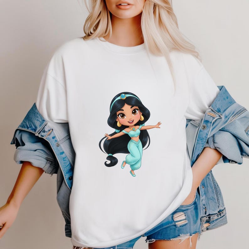 Puede incluir: Camiseta blanca con una ilustraci&oacute;n de dibujos animados de una princesa con un atuendo turquesa, pelo largo negro, una diadema azul y pendientes dorados. La princesa est&aacute; en una pose de baile. La camisa la lleva una persona con una chaqueta vaquera.