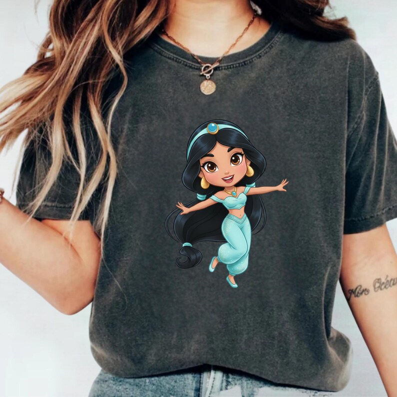 Puede incluir: Camiseta gris oscuro con una ilustraci&oacute;n de dibujos animados de la princesa Jasmine con un atuendo turquesa y pelo largo negro. La princesa sonr&iacute;e con los brazos extendidos. La camiseta es de estilo casual.