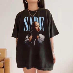 Op de afbeelding: Zwart T-shirt met een portret van Sade Adu en de naam "SADE" in lichtblauw boven de afbeelding. Sade draagt een zwarte handschoen en een wit polkadotshirt. Een kledingstuk met een muziekthema.
