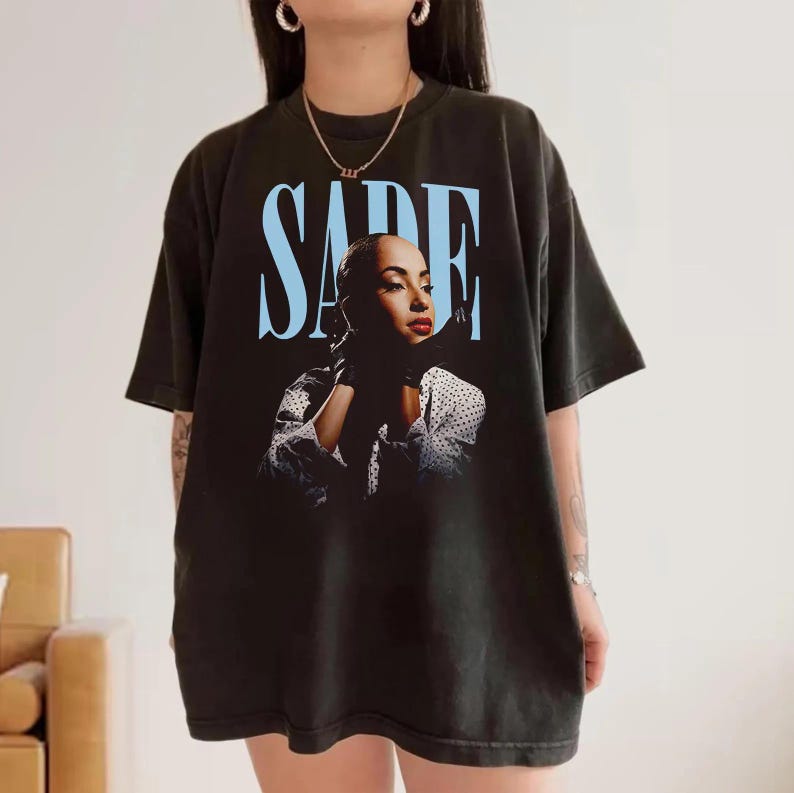 Op de afbeelding: Zwart T-shirt met een portret van Sade Adu en de naam "SADE" in lichtblauw boven de afbeelding. Sade draagt een zwarte handschoen en een wit polkadotshirt. Een kledingstuk met een muziekthema.