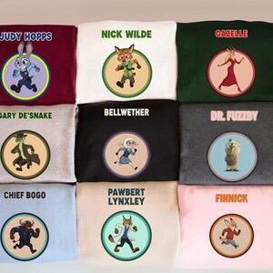 May include: A collection of nine sweatshirts in various colors, each featuring a circular graphic of a character from the animated film "Zootopia." The sweatshirts display character names like "Judy Hopps," "Nick Wilde," and "Gazelle."