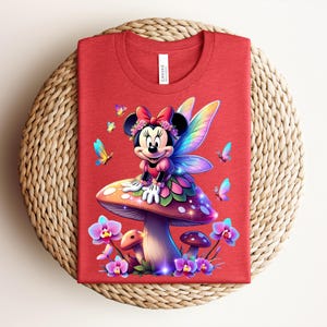 May include: Red t-shirt featuring a colorful illustration of a cartoon character with fairy wings, sitting on a mushroom. The design includes butterflies and flowers. The shirt has a round neck and a tag that says "Canvas".