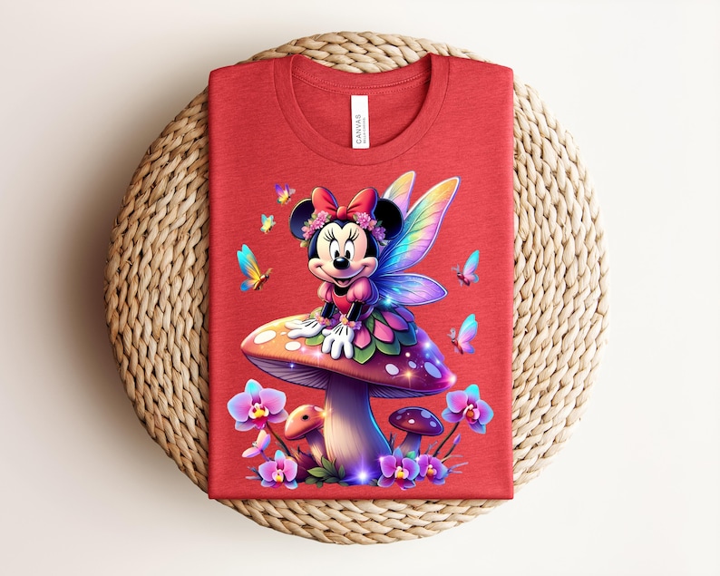 May include: Red t-shirt featuring a colorful illustration of a cartoon character with fairy wings, sitting on a mushroom. The design includes butterflies and flowers. The shirt has a round neck and a tag that says "Canvas".