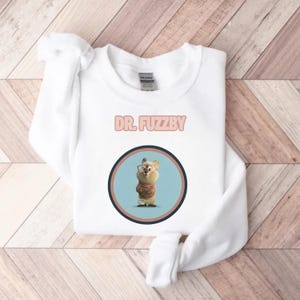 May include: White sweatshirt with the text "DR. FUZZBY" in pink. The sweatshirt features a circular graphic of a cartoon character wearing glasses and a sweater. The graphic is set against a light blue background.