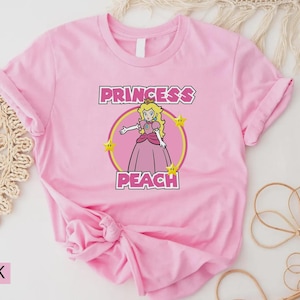 Może przedstawiać: R&oacute;żowa koszulka z grafiką Princess Peach. Projekt zawiera napis "PRINCESS PEACH" nad i pod ilustracją postaci w ż&oacute;łtym okręgu.