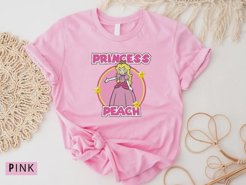 Może przedstawiać: R&oacute;żowa koszulka z grafiką Princess Peach. Projekt zawiera napis "PRINCESS PEACH" nad i pod ilustracją postaci w ż&oacute;łtym okręgu.