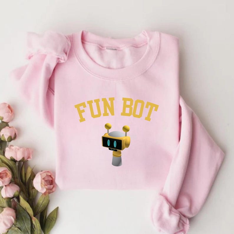 May include: A light pink sweatshirt with the text "FUN BOT" in gold lettering. Below the text is a cartoon robot with a yellow head, black screen, and gray body. The sweatshirt has long sleeves and a crew neck.