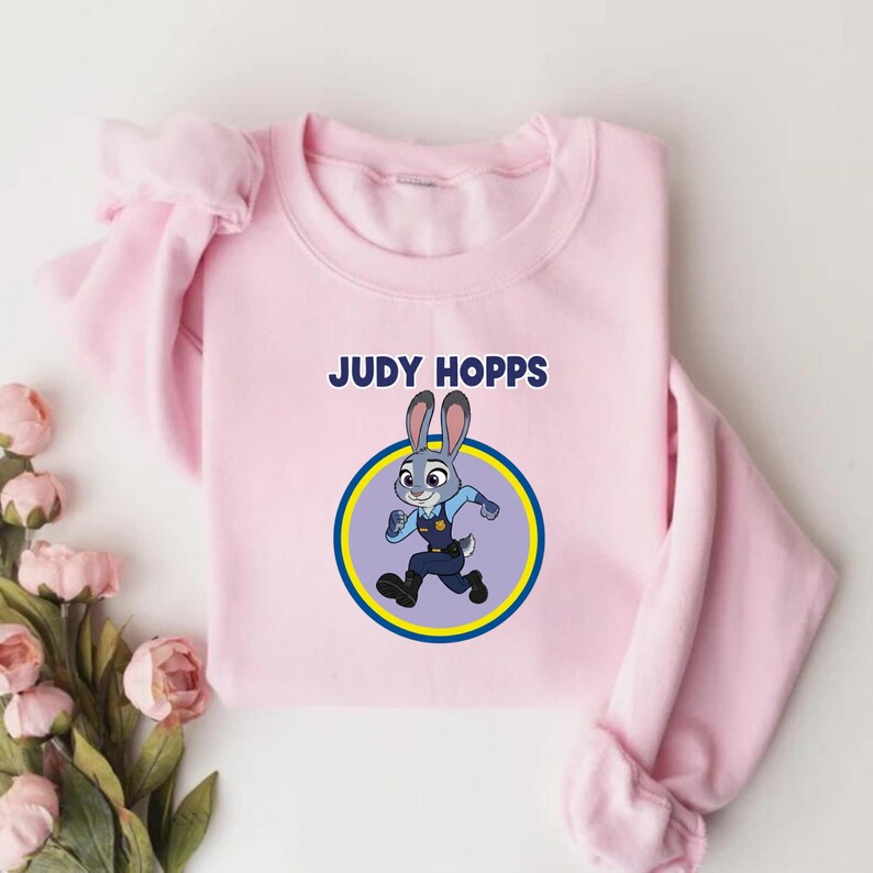 以下が含まれることがあります： 「JUDY HOPPS」の文字と、走っているウサギのキャラクター、ジュディ・ホップスの漫画のイメージが描かれた、薄いピンク色のスウェットシャツ。 黄色と青の円の中に画像があります。