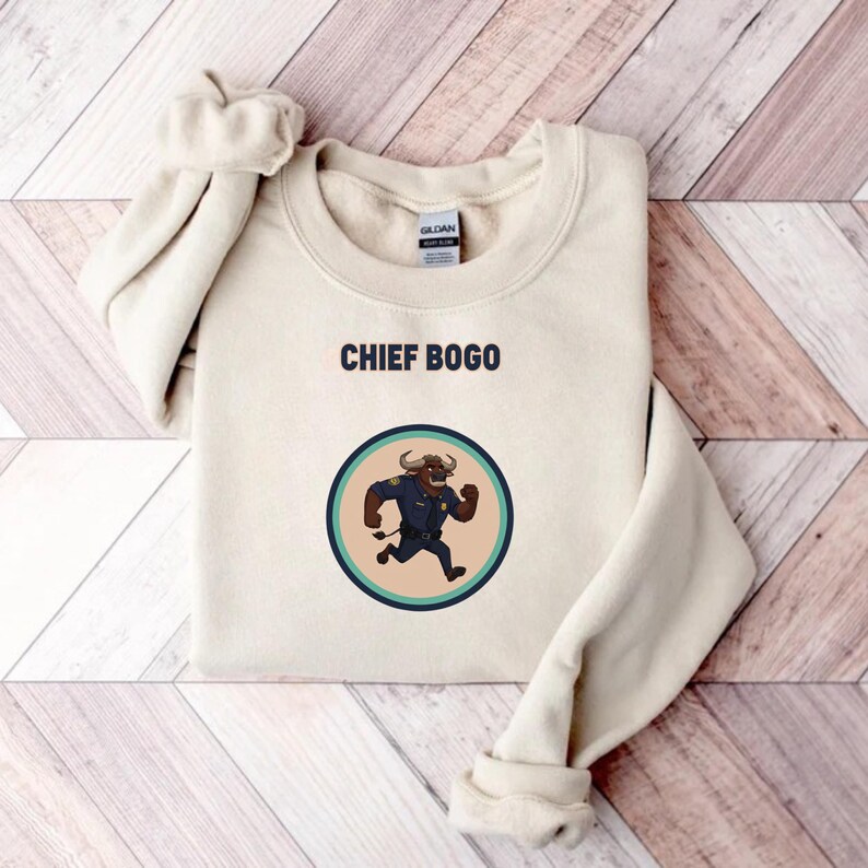 May include: A cream-colored sweatshirt with the text "CHIEF BOGO" above a circular graphic. The graphic features a cartoon bull in a police uniform running. The sweatshirt is folded on a wooden surface.