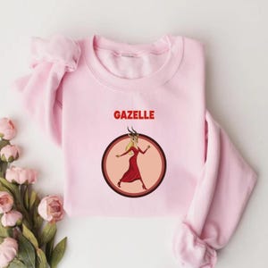 May include: A pale pink sweatshirt with the word "GAZELLE" in red above a circular graphic. The graphic shows a cartoon gazelle woman in a red dress and heels. The gazelle is inside a brown circle with a black outline.
